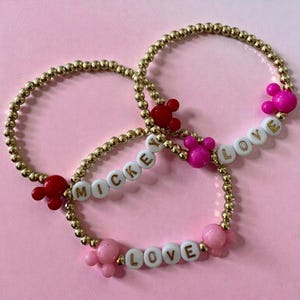 May include: Three gold beaded bracelets with white letter beads spelling "MICKEY" and "LOVE". Each bracelet features a Mickey Mouse head charm in red or pink. The bracelets are displayed on a pink background.