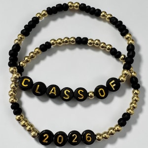 May include: Two beaded bracelets. One bracelet features the text "CLASS OF" in black beads with gold lettering. The other bracelet has the text "2026" in black beads with gold lettering. The bracelets are made of black and gold beads.