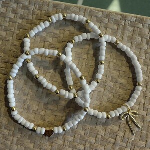 May include: Three white beaded bracelets with gold-colored accents and a gold bow charm. One bracelet has a small wooden heart charm. The bracelets are arranged on a woven surface.