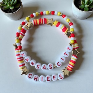 May include: Two beaded bracelets with the words "Kansas City Chiefs". The bracelets feature red, yellow, and white beads, gold star charms, and gold accent beads. The bracelets are on a white surface.