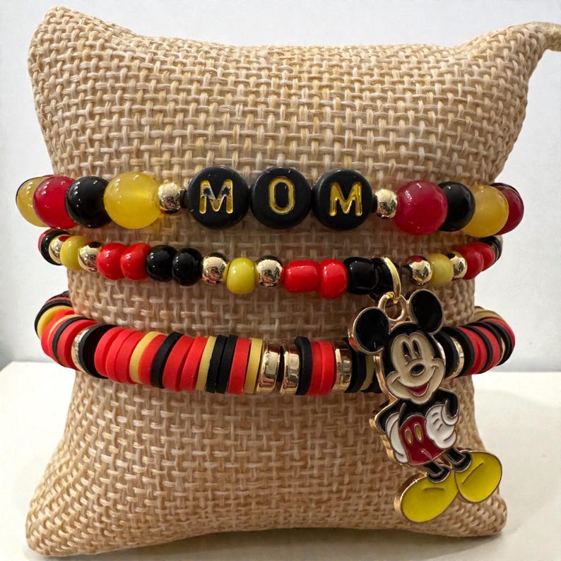 Puede incluir: Conjunto de cuatro pulseras con cuentas rojas, negras, amarillas y doradas. Una pulsera deletrea "MOM" en letras negras. Un dije de Mickey Mouse cuelga de una pulsera. Las pulseras se exhiben sobre un coj&iacute;n beige.