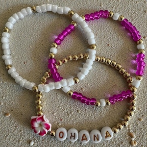 May include: Three beaded bracelets. One bracelet is white with gold beads, another is fuchsia with gold beads, and the third is gold with the word "OHANA" spelled out in white beads. A pink and white flower charm is attached.