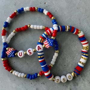 May include: Three patriotic bracelets in red, white, and blue. One bracelet spells "USA" with letter beads. Another features a star charm with the American flag. Gold-colored beads accent the designs. The bracelets are on a gray surface.