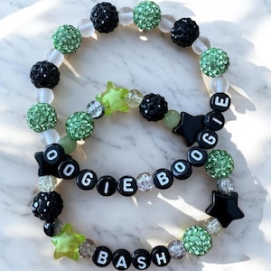 May include: Two beaded bracelets with the words "Oogie Boogie Bash". The bracelets feature black, green, and clear beads, along with star-shaped and round beads. The letters are white on black beads.