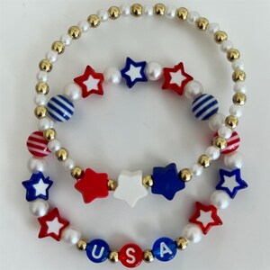 May include: Two beaded bracelets featuring a patriotic theme. One bracelet consists of white pearl-like beads, gold metallic beads, blue and white striped beads, and red, white, and blue star-shaped beads. The second bracelet includes white pearl-like beads, gold metallic beads, red and white striped beads, red, white, and blue star-shaped beads, and blue beads with the white letters U, S, and A.