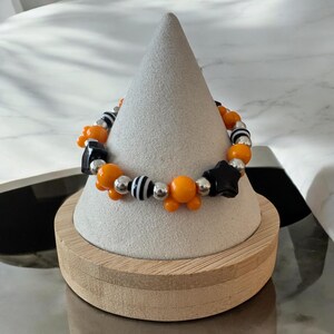 May include: A beaded bracelet featuring orange, black, silver, and white beads. The bracelet includes orange Mickey Mouse-shaped beads, black star-shaped beads, and silver accent beads. The bracelet is displayed on a gray cone-shaped stand.