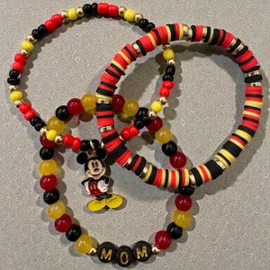 Puede incluir: Tres pulseras de cuentas rojas, negras, amarillas y doradas. Una pulsera presenta un dije de Mickey Mouse, y otra deletrea "MOM" en letras negras. Las pulseras est&aacute;n dispuestas en un patr&oacute;n circular.