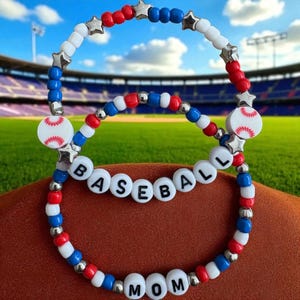 May include: Two beaded bracelets with a baseball theme. One bracelet spells out "BASEBALL" and the other says "MOM". The bracelets are red, white, and blue with baseball and star charms. The background is a baseball field.
