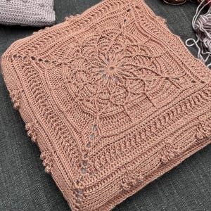 May include: A square, hand-crocheted cushion in a dusty rose color. The cushion features a central floral design and intricate border details. The yarn appears to be a soft, natural fiber. A second, similar cushion is partially visible.