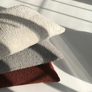 Può includere: Tre cuscini decorativi strutturati impilati. Il cuscino superiore è color crema, il cuscino centrale è grigio e il cuscino inferiore è color ruggine scuro. I cuscini sono disposti su una superficie bianca con ombre.