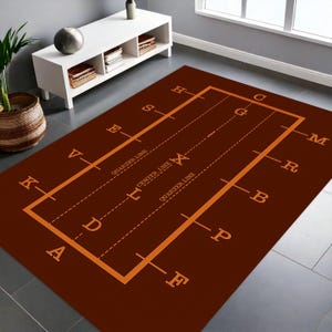 Könnte beinhalten: Ein rechteckiger, brauner Teppich mit einem orangefarbenen Footballfeld-Design. Der Teppich zeigt Yard-Linien, Viertellinien und eine Mittellinie, mit Buchstaben, die den Umfang des Feldes markieren. Der Teppich liegt auf einem gefliesten Boden.