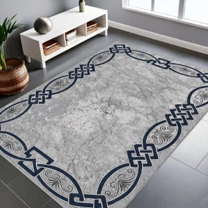 May include: A rectangular gray rug with a distressed texture and a dark blue border. The border features a repeating pattern of interlocking shapes and floral motifs. The rug is placed on a gray tiled floor.