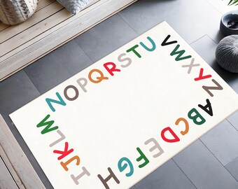 May include: A rectangular off-white rug with the alphabet in various colors forming a border. The letters are in a playful font, with a white center. The rug is on a dark gray floor.