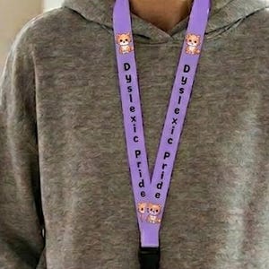 Può includere: Un cordino viola con la scritta "Dyslexic Pride" e personaggi dei cartoni animati è indossato al collo. Il cordino porta un badge identificativo trasparente con la scritta "Staff". La persona indossa una felpa grigia con cappuccio e pantaloni beige.