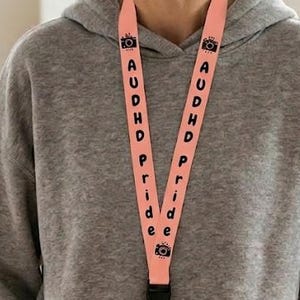 Könnte beinhalten: Ein pfirsichfarbenes Schlüsselband mit dem Text "AUDHD PRIDE" in Schwarz und Kamera-Symbolen. Es hält einen transparenten Ausweis. Die Person trägt einen grauen Kapuzenpullover und eine beige Hose.