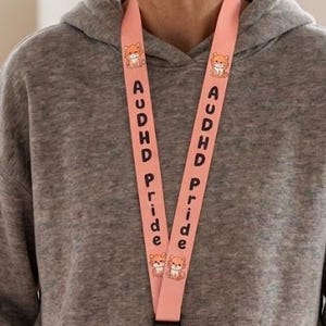 Può includere: Un cordino rosa con la scritta "AUDHD Pride" e un disegno di animale dei cartoni animati. Il cordino porta un tesserino bianco con una foto e la scritta "STAFF 0014". La persona indossa una felpa con cappuccio grigia e pantaloni rosa.