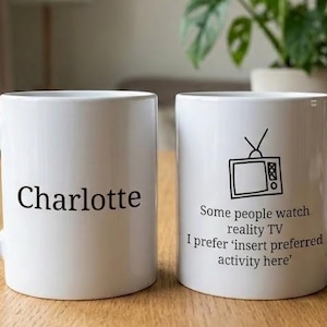 May include: Two white ceramic mugs on a wooden surface. One mug has the name "Charlotte" printed in black. The other mug features a retro TV graphic and the text: "Some people watch reality TV I prefer 'insert preferred activity here'."