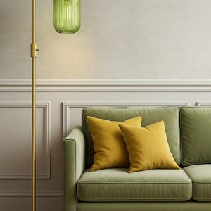 May include: A floor lamp with a slender brass stand and a ribbed, translucent green glass shade, positioned next to a sage green sofa featuring two square mustard yellow throw pillows. The lamp has a circular base and a vertical adjustable neck.