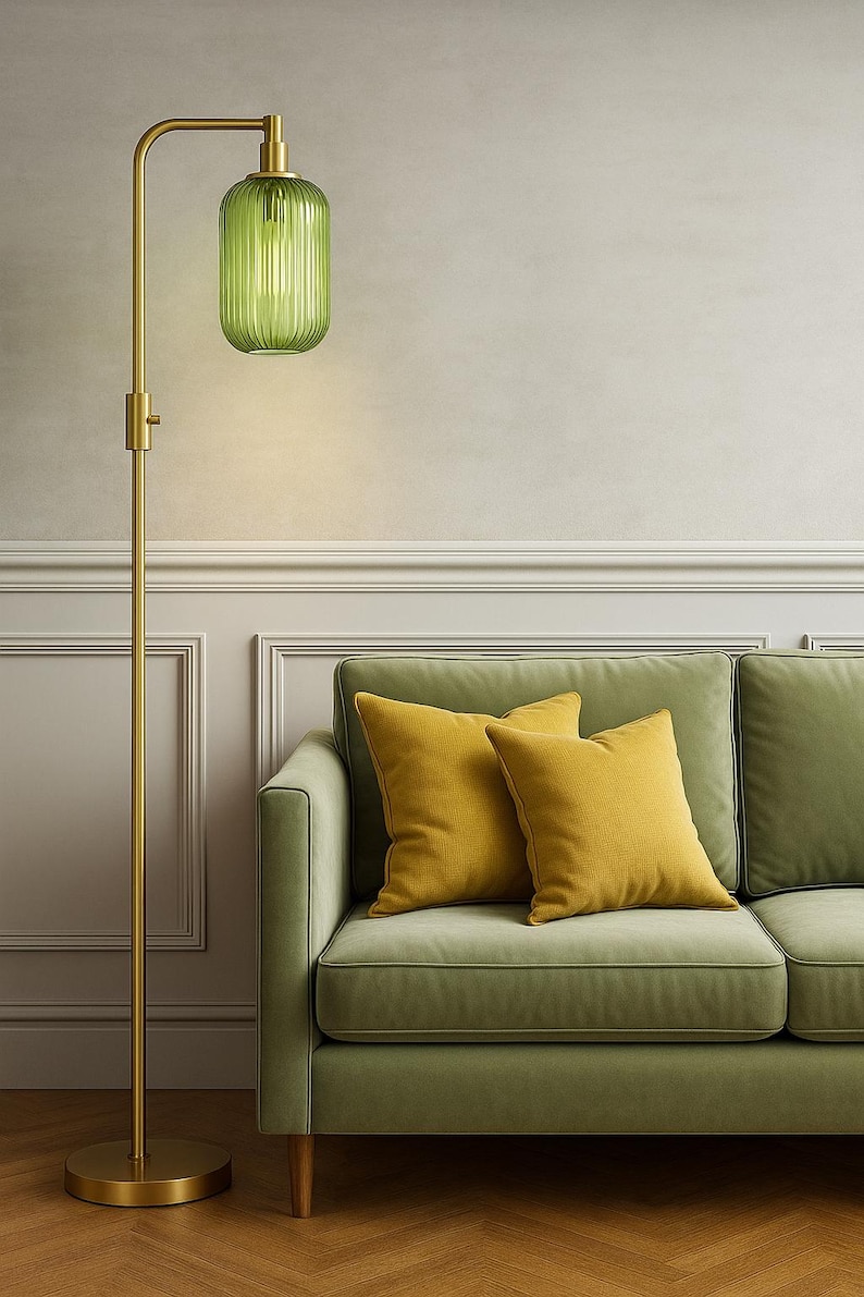 May include: A floor lamp with a slender brass stand and a ribbed, translucent green glass shade, positioned next to a sage green sofa featuring two square mustard yellow throw pillows. The lamp has a circular base and a vertical adjustable neck.