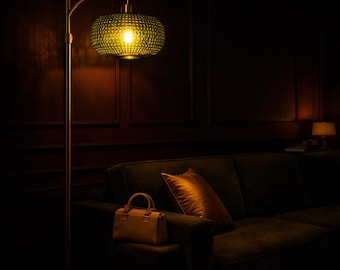 May include: A gold floor lamp with a textured green glass shade illuminates a dark room. A brown leather handbag and a gold pillow rest on a dark green sofa. The lamp's warm light creates a cozy atmosphere.