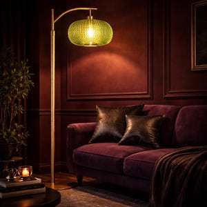 May include: A gold floor lamp with a green, textured glass shade illuminates a room with a burgundy sofa and pillows. The lamp's curved arm extends over the sofa, casting a warm glow. The room has a cozy, inviting atmosphere.
