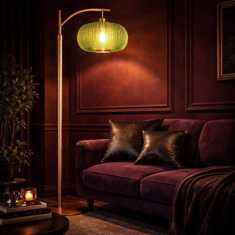 May include: A gold floor lamp with a green, textured glass shade illuminates a room with a burgundy sofa and pillows. The lamp's curved arm extends over the sofa, casting a warm glow. The room has a cozy, inviting atmosphere.