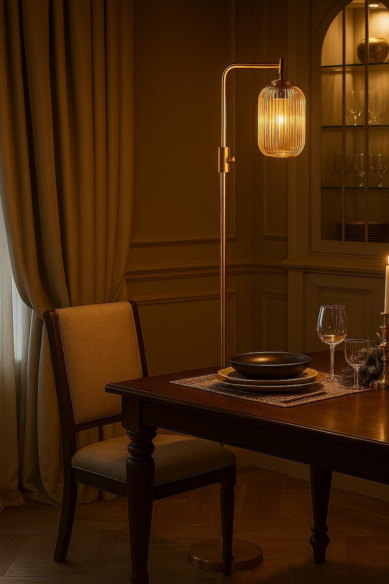 May include: A tall floor lamp with a slender metallic stand and a cylindrical, vertically ribbed glass shade that emits a warm glow. The lamp is positioned next to a wooden dining table set with stacked plates, a bowl, and glassware.