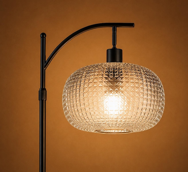 May include: A black metal floor lamp with a curved arm supporting a textured, clear glass shade. The shade has a diamond pattern and houses a bright light bulb. The lamp is a stylish lighting fixture.