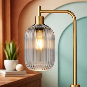 May include: A table lamp featuring a brushed gold metal frame with an L-shaped arm and a cylindrical, vertically ribbed, translucent gray glass shade. The lamp contains a warm-toned light bulb that illuminates the interior of the glass shade.