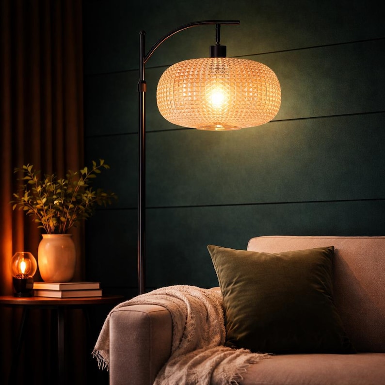 May include: A modern floor lamp with a textured, amber-colored glass shade. The lamp has a black metal frame and a curved arm. The lamp is positioned near a sofa with a green pillow and a dark green wall.