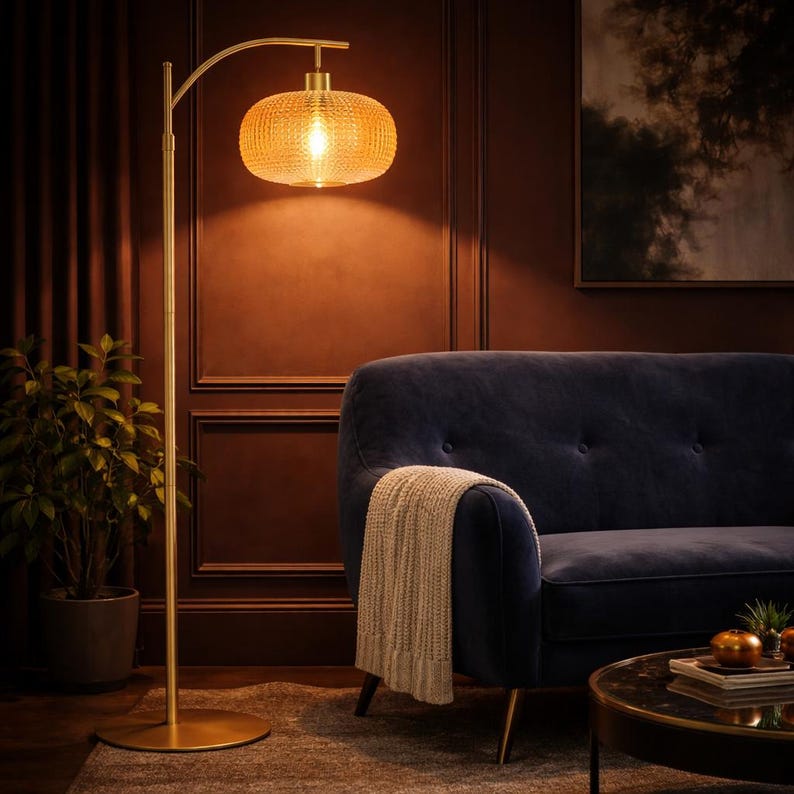 May include: A gold-toned floor lamp with a textured, amber-colored glass shade illuminates a living room. The lamp has a curved arm and a round base. A navy blue velvet sofa and a potted plant are also visible.