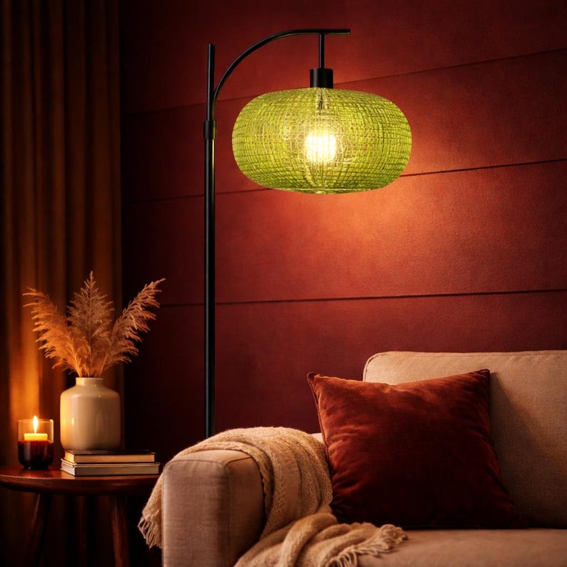 May include: A modern floor lamp with a woven green lampshade. The black metal frame arches over a beige sofa with a rust-colored pillow. A small table holds a vase with dried plants and a lit candle.
