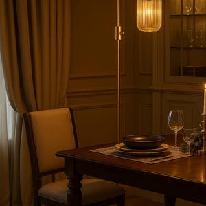 May include: A tall floor lamp with a slender metallic stand and a cylindrical, vertically ribbed glass shade that emits a warm glow. The lamp is positioned next to a wooden dining table set with stacked plates, a bowl, and glassware.