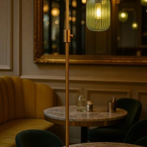 May include: A floor lamp with a slender, matte gold metal stand and a single, vertically ribbed, translucent green glass shade. The lamp features a simple cylindrical design with a warm, glowing light bulb visible inside the glass fixture.
