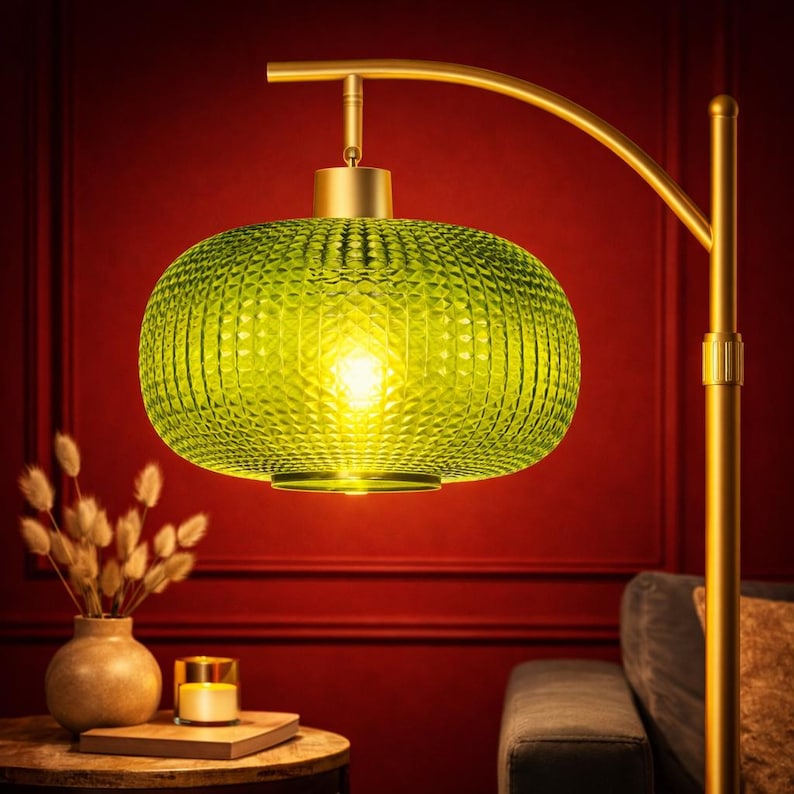 May include: A modern floor lamp with a gold-colored frame and a large, textured, green glass lampshade. The lampshade is oval-shaped and emits a warm, yellow light. The lamp is set against a red wall.
