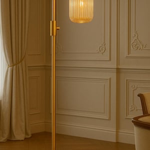 May include: A tall floor lamp with a brushed gold metal finish, featuring a slender vertical pole, a circular base, and an adjustable arm. The light source is enclosed in a cylindrical, vertically ribbed amber glass shade that emits a warm glow.