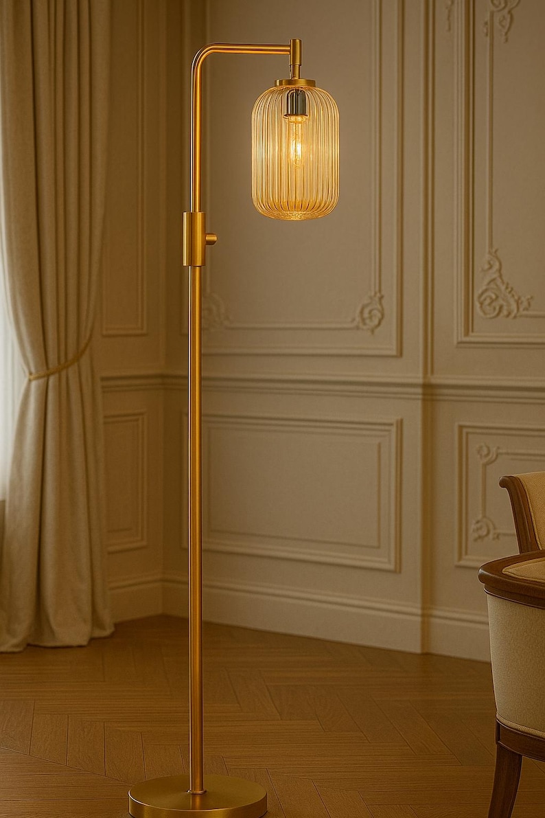 May include: A tall floor lamp with a brushed gold metal finish, featuring a slender vertical pole, a circular base, and an adjustable arm. The light source is enclosed in a cylindrical, vertically ribbed amber glass shade that emits a warm glow.