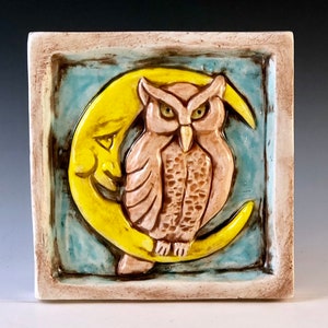 May include: A ceramic tile featuring a raised design of an owl perched on a crescent moon. The owl is brown with yellow eyes and the moon is yellow. The tile is framed in a light brown border.