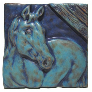 May include: A ceramic tile with a blue and brown glaze depicting a horse's head in relief. The horse's head is facing to the left and the background is a mottled blue.