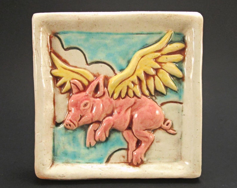 May include: A ceramic tile featuring a pink pig with yellow wings flying in a blue sky with white clouds.
