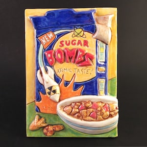 May include: A ceramic tile depicting a box of "New Sugar Bombs" cereal with an atomic taste. The box is blue with red and yellow lettering. The tile also features a bowl of cereal and a few pieces of cereal scattered around the bowl.