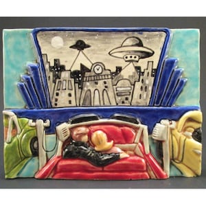 May include: A ceramic tile depicting a drive-in movie theater scene. Two people are sitting in a red car, watching a movie on a large screen. The movie features a city skyline with flying saucers in the night sky.