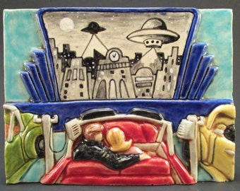 May include: A ceramic tile depicting a drive-in movie theater scene. Two people are sitting in a red car, watching a movie on a large screen. The movie features a city skyline with flying saucers in the night sky.