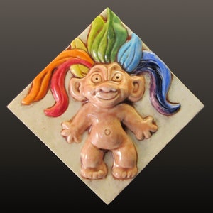 May include: A ceramic tile with a raised relief sculpture of a troll with rainbow colored hair. The troll is smiling and has a light pink skin tone.