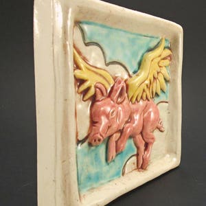 May include: A ceramic tile featuring a hand-painted design of a pink pig with yellow wings flying through the clouds. The tile has a white background with blue and green accents.