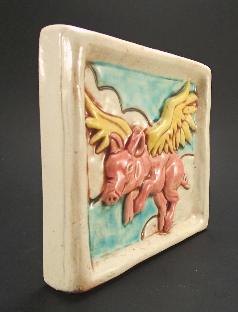 May include: A ceramic tile featuring a hand-painted design of a pink pig with yellow wings flying through the clouds. The tile has a white background with blue and green accents.