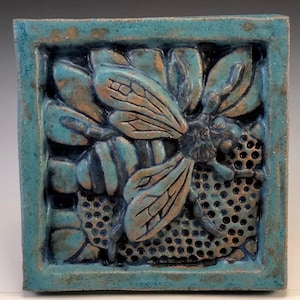 Może przedstawiać: Płytka ceramiczna z niebieskim i brązowym szkliwem przedstawiająca pszczołę w plastrze miodu. Pszczoła jest w reliefie i ma szczegółowe skrzydła i nogi.