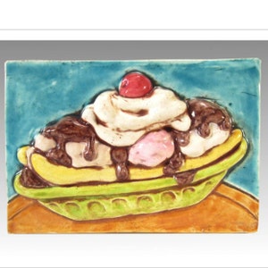 May include: A ceramic tile with a raised image of a banana split. The banana split is topped with whipped cream, chocolate sauce, a cherry, and two scoops of ice cream. The tile has a blue background and is set on a brown surface.