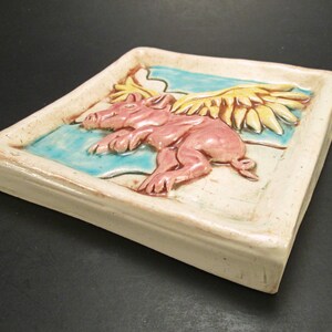 May include: A ceramic tile featuring a pink pig with yellow wings flying over a blue and white background.