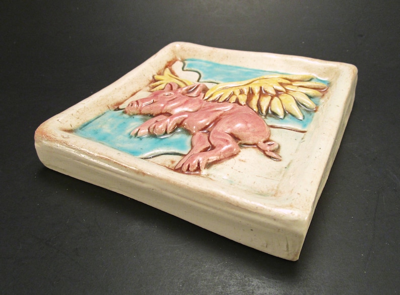 May include: A ceramic tile featuring a pink pig with yellow wings flying over a blue and white background.